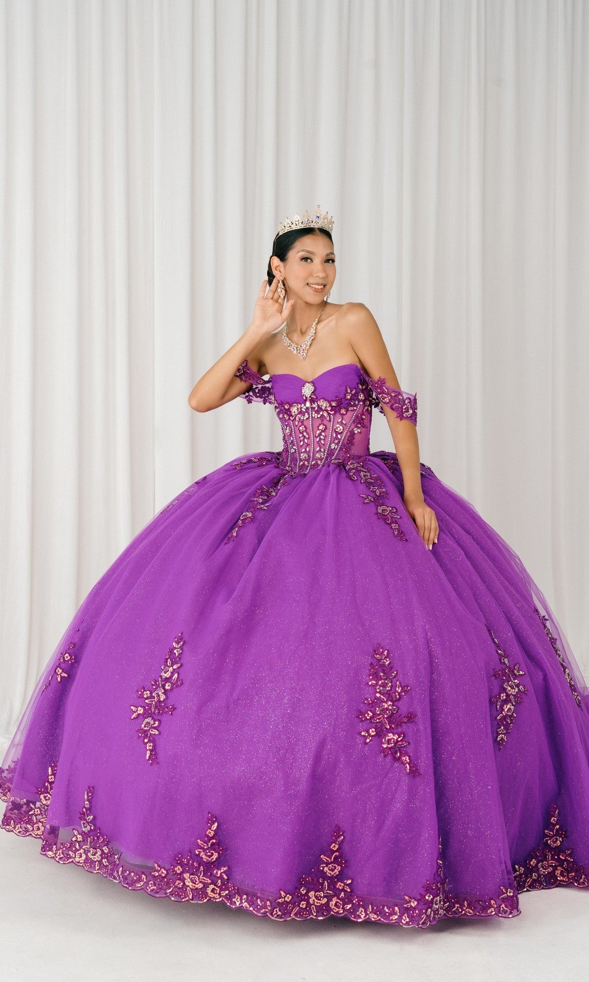 A woman in a magenta Quinceanera Dress 8405JQ by Cinderella Couture with gold accents and a tiara poses in front of white curtains, holding one hand near her ear.